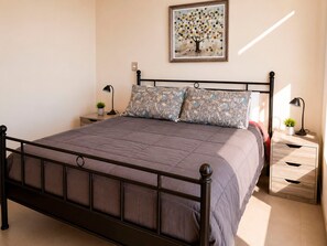 A neatly made bed with a grey quilt, two bedside tables with lamps, a small potted plant, and a framed artwork on the wall.