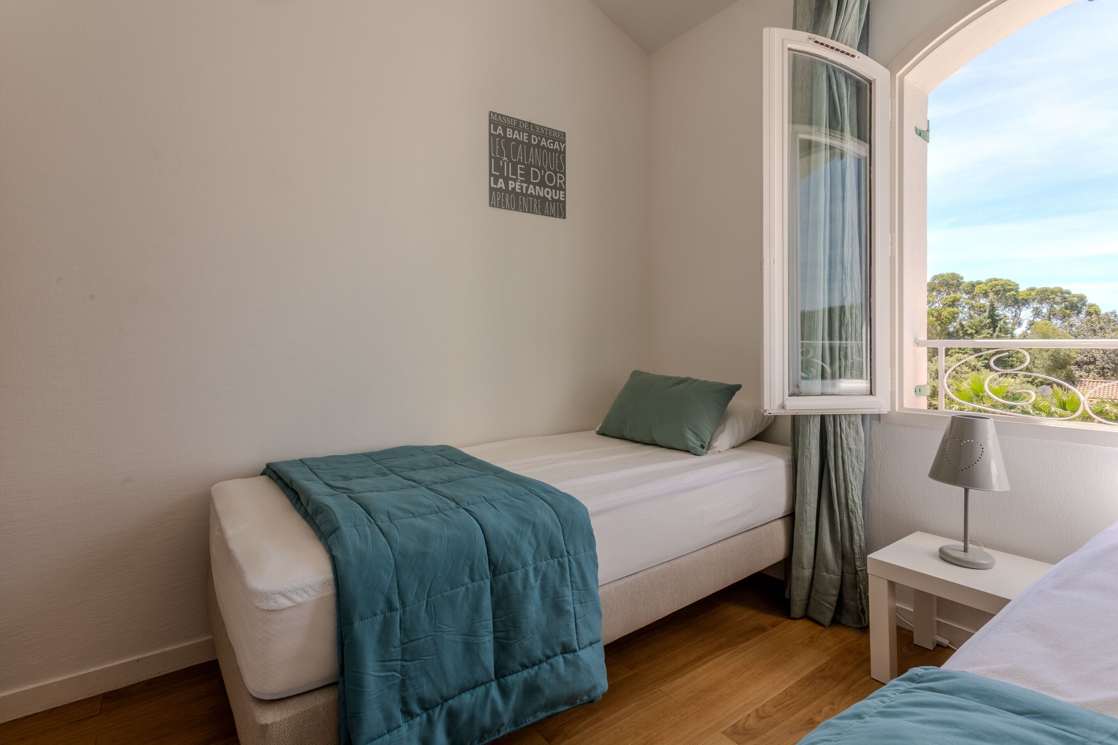 Une chambre avec un lit simple, une fenêtre avec des rideaux, une table de chevet avec une lampe et un mur orné d’un panneau avec une inscription.
