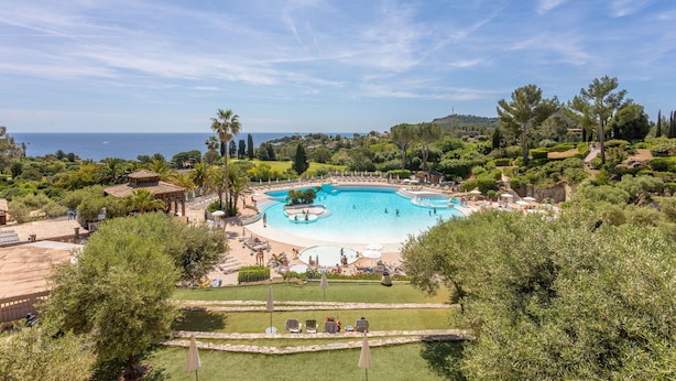 A resort with a large swimming pool, lush greenery, and a view of the sea.
