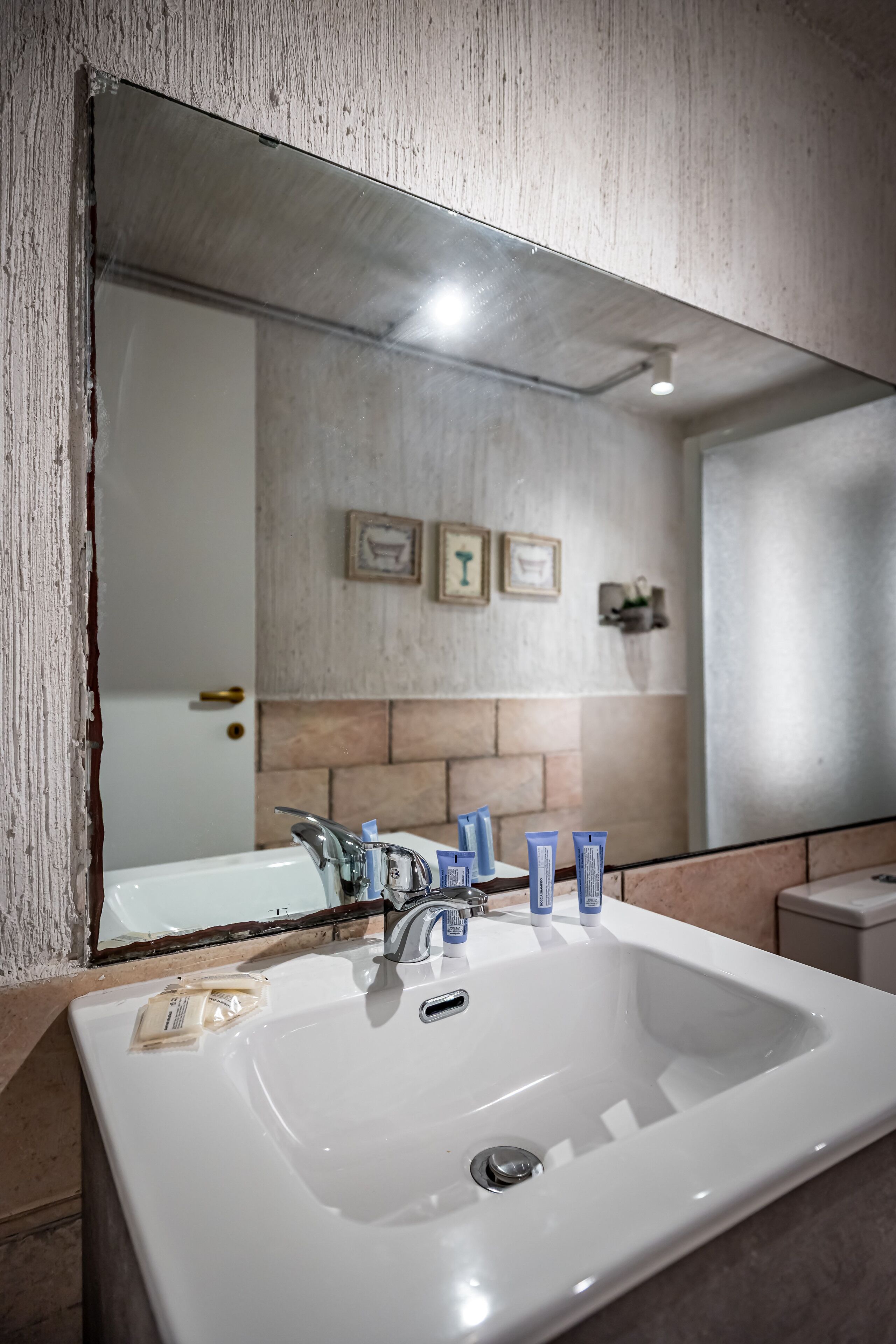 A bathroom with a large mirror, white sink, and two faucets. There are toiletries on the sink and a towel rack on the wall.