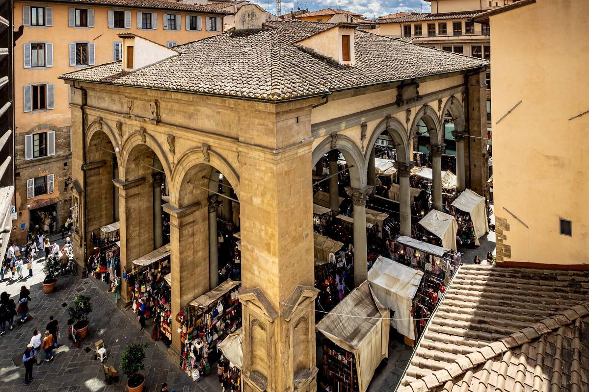 A bustling square with historical architecture, including arches and a tower.