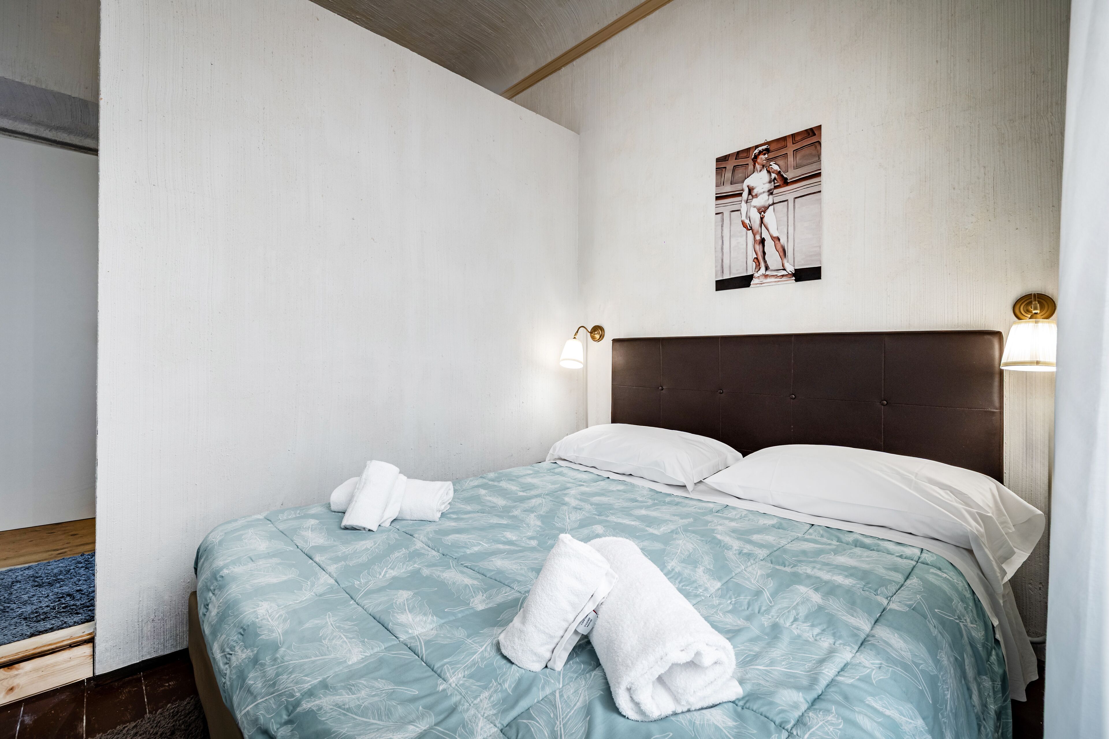 A neatly made bed with a blue floral bedspread and white pillows, two white towels on the bed, and a framed artwork above the bed.