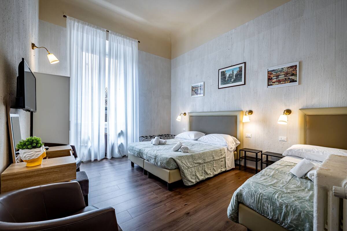 A hotel room with a bed, bedside tables, a desk with a chair, a plant, and a window with curtains.
