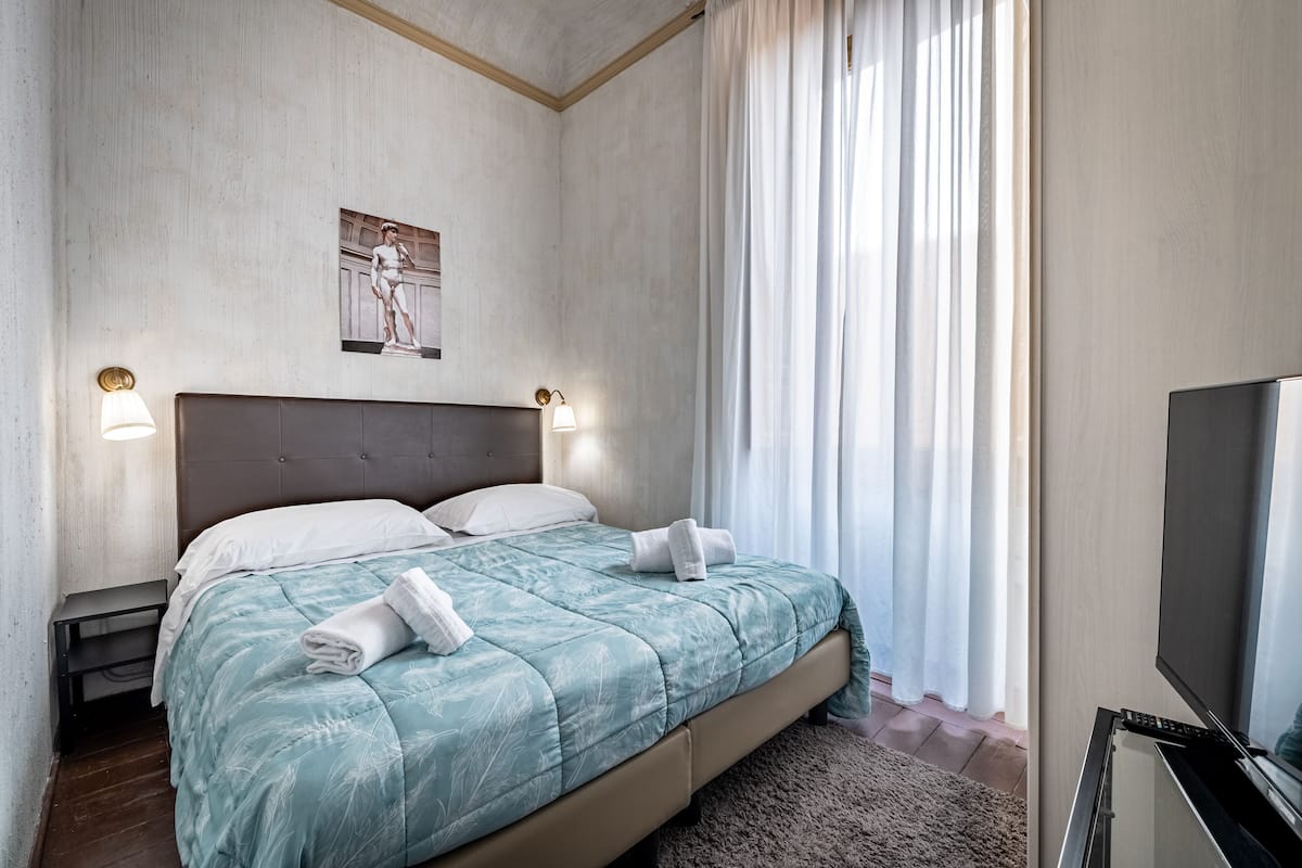 A neatly made bed with a teal comforter and white pillows, a wall-mounted artwork, a television on a stand, and a large window with sheer curtains.