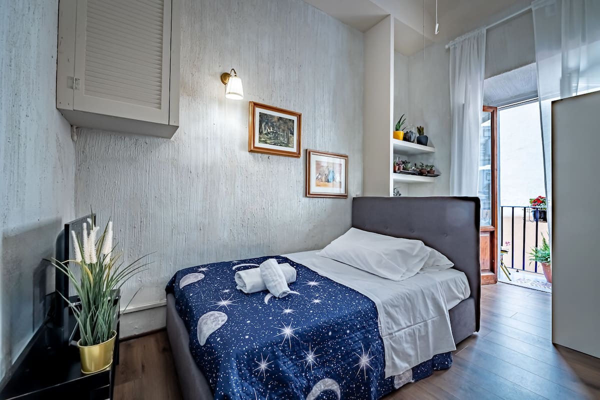 A modern bedroom with a bed, a nightstand, a wall-mounted lamp, framed pictures, and a balcony with plants.