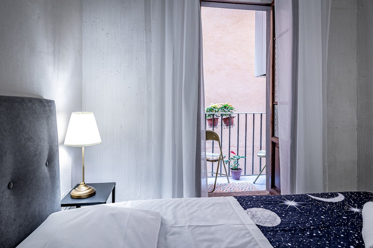 A hotel room with a bed, a nightstand with a lamp, a view of a balcony with potted plants.