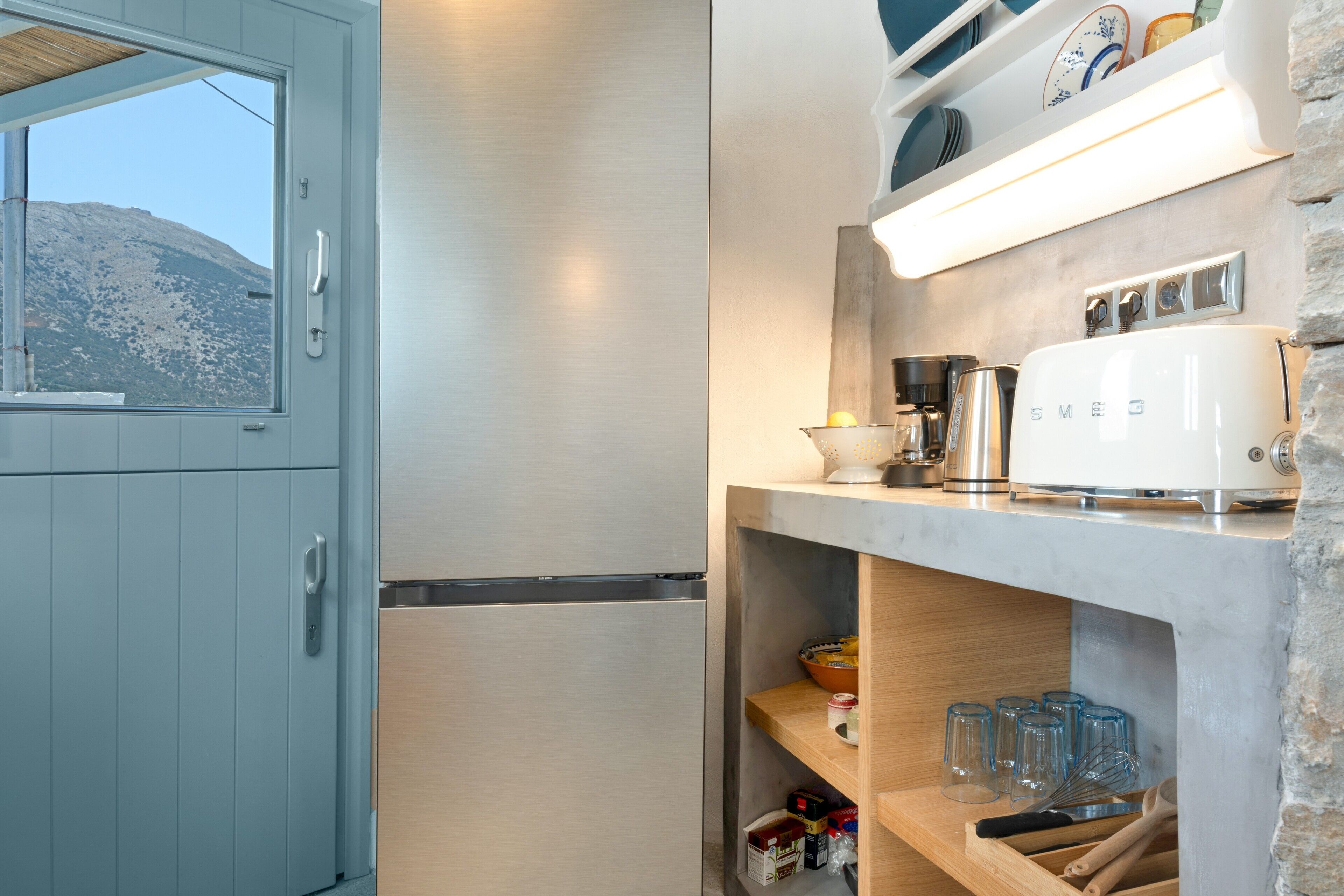 Una cocina compacta con un refrigerador de acero inoxidable, una tetera eléctrica blanca y un tostador sobre una repisa.