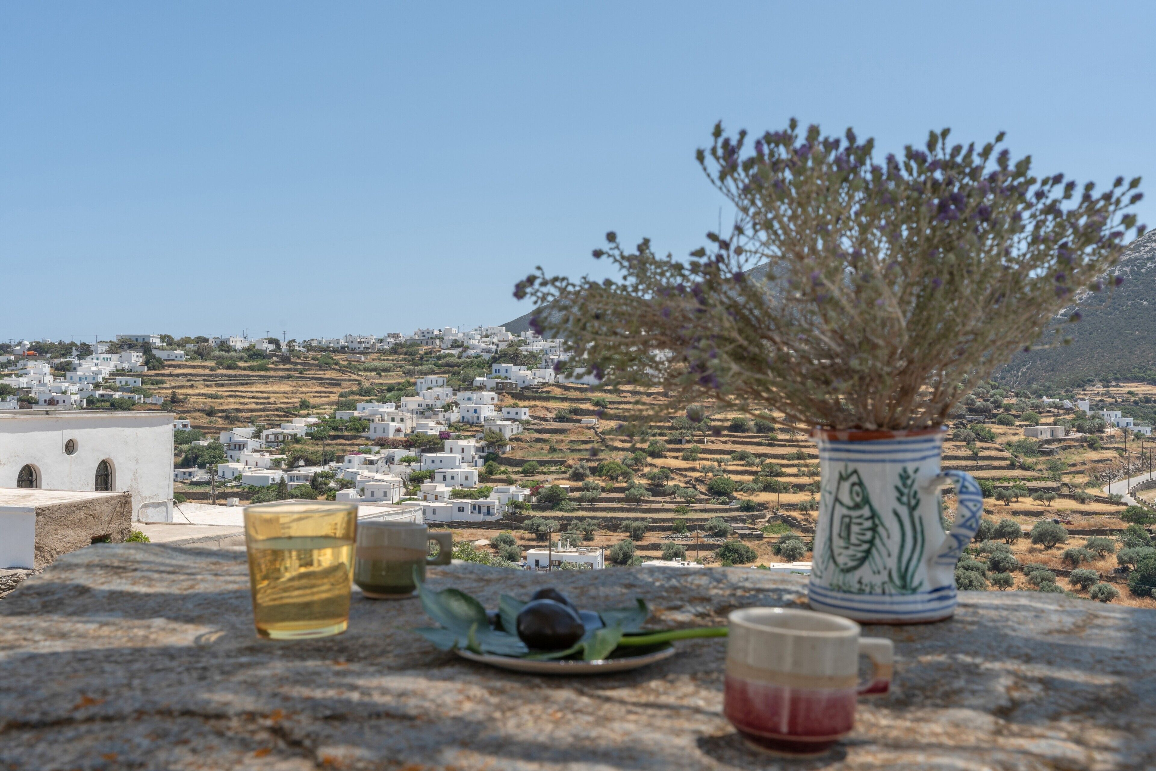 A stone table with a vase, a plate with olives, and two glasses, overlooking a coastal village with white buildings.
