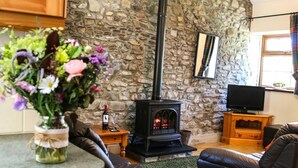 A cozy living room with a stone wall, a fireplace, leather sofas, a television, and a vase of flowers. - Y Dderwen, Aberaeron (Llanarth)