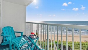 A balcony with a turquoise Adirondack chair and a small table with a bottle and a glass. The balcony overlooks a beach with the ocean and a clear sky.