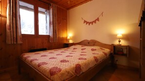 A wooden room with a bed, two bedside lamps, a window with curtains, and a heart-shaped wall decoration.