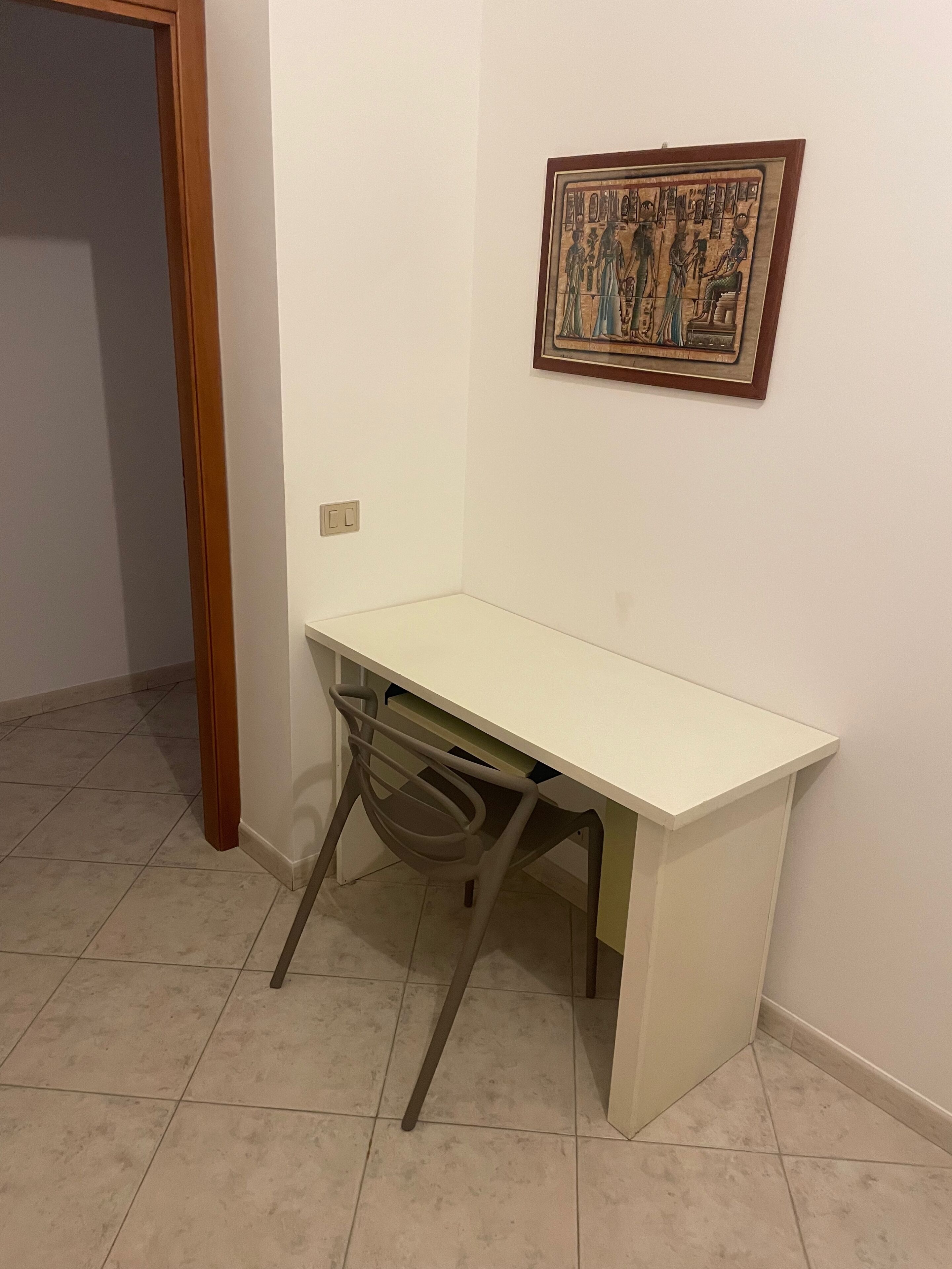 Una habitación con un escritorio blanco, una silla, un cuadro en la pared y una puerta.