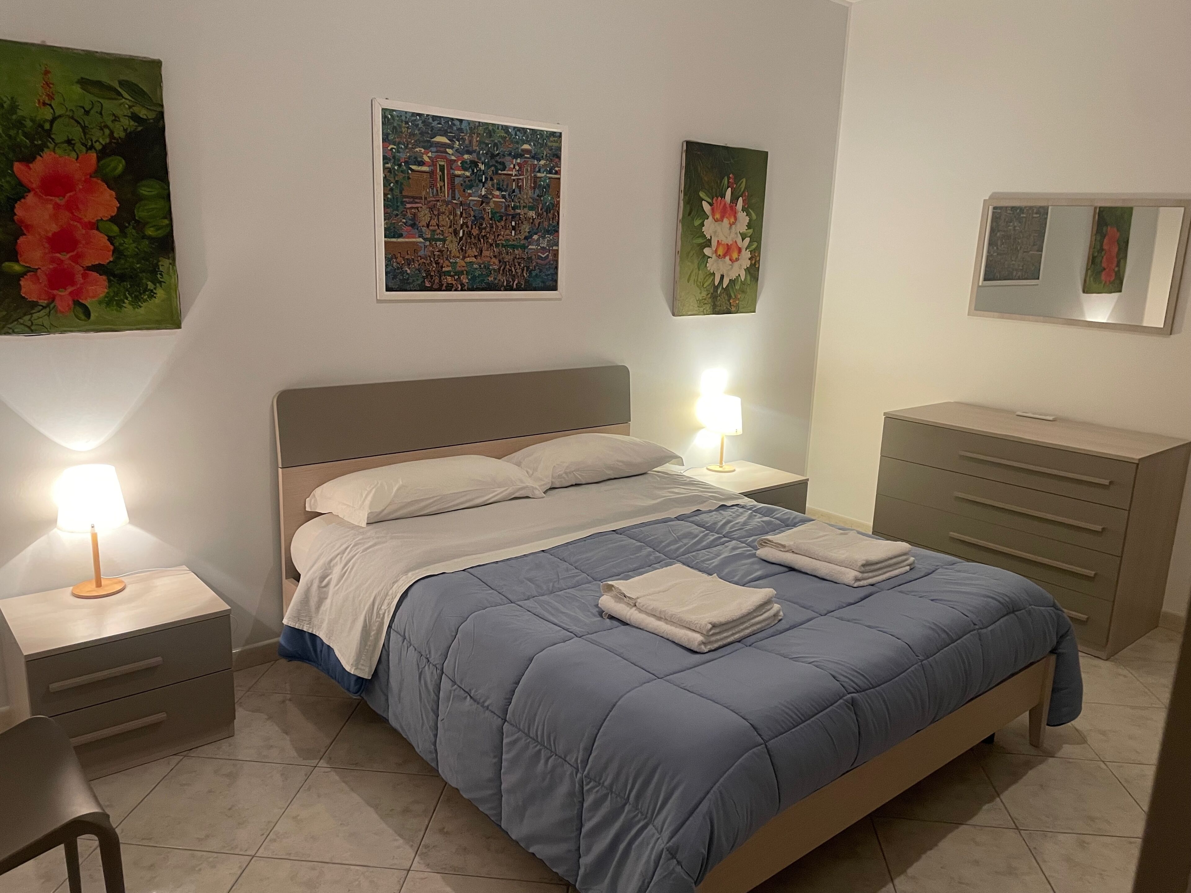 A neatly made bed with a blue quilt, two bedside lamps, a dresser, and wall art.