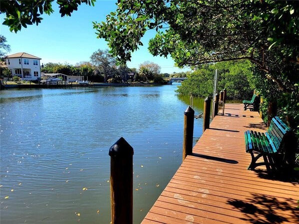 A wooden dock extending over calm water, with a bench and a pathway leading to a house in the background.
