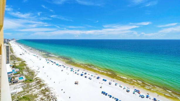 Una playa con arena blanca y agua turquesa cristalina. Hay sombrillas azules y sillas de playa a lo largo de la orilla.