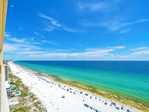 Ein Strand mit weißem Sand und klarem, türkisfarbenem Wasser. Entlang der Küste befinden sich blaue Sonnenschirme und Liegestühle.