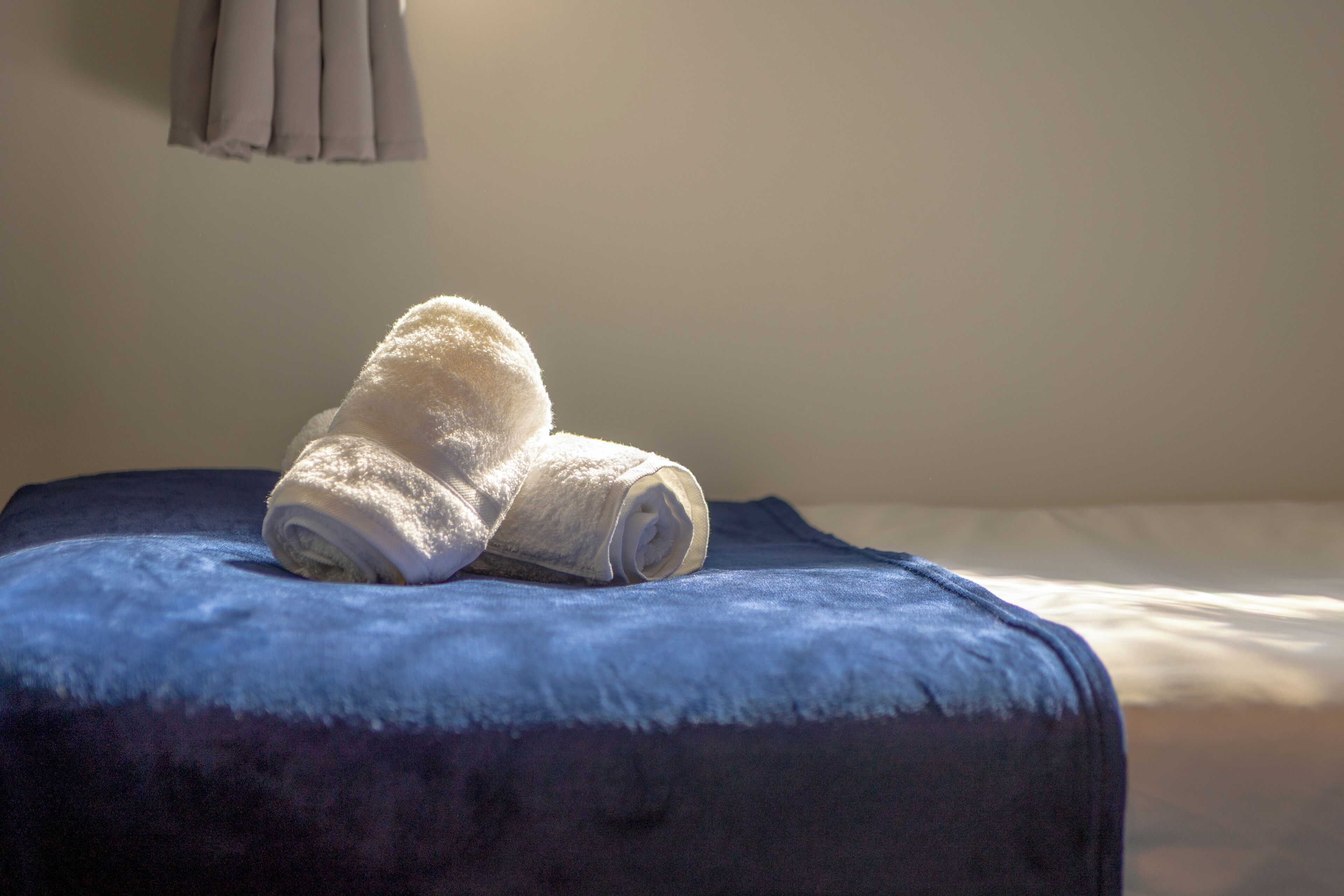A neatly made bed with a blue bedspread and a rolled white towel on top.