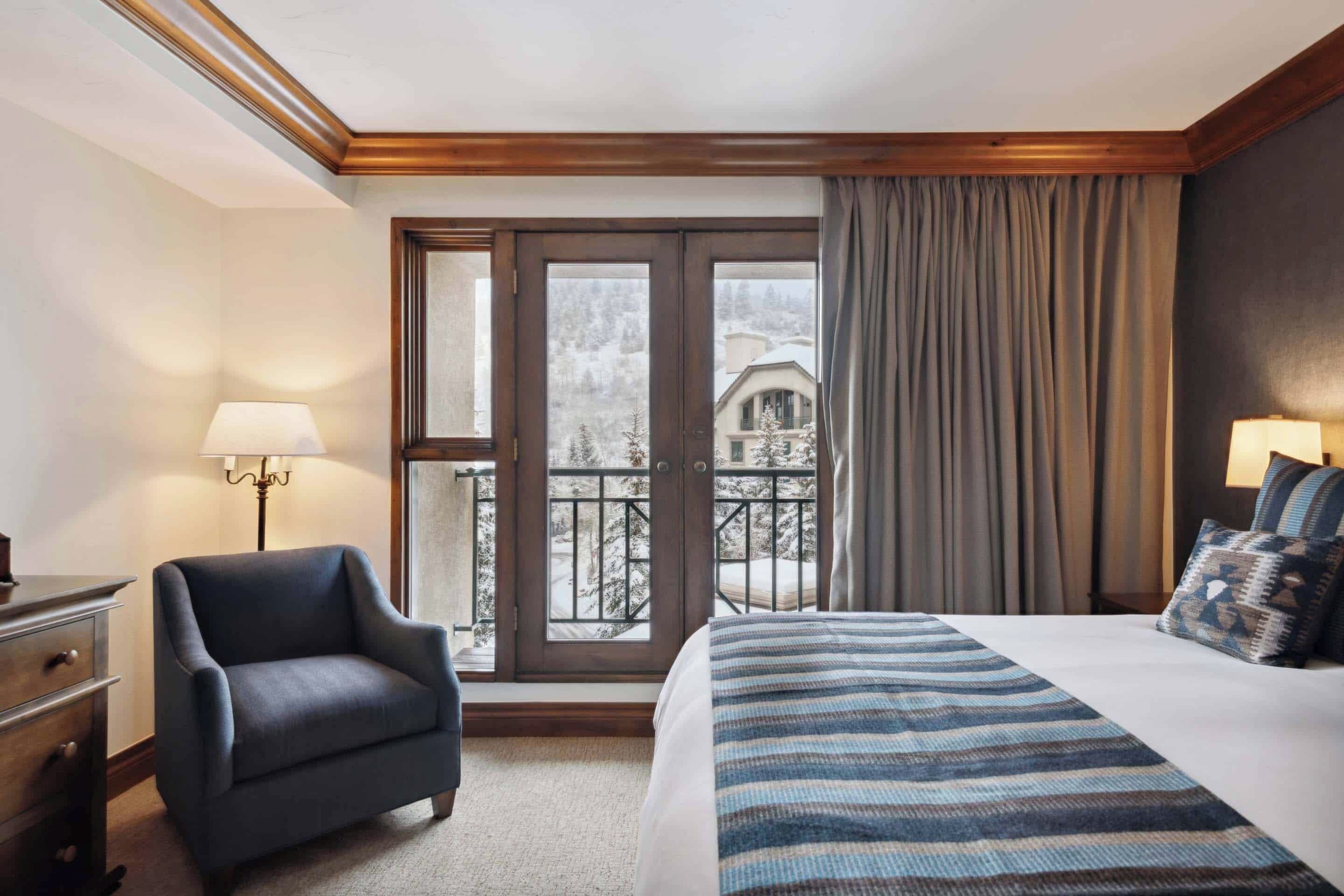 Habitación de hotel con cama, silla, escritorio, lámpara y vista a un paisaje nevado a través de un ventanal grande.