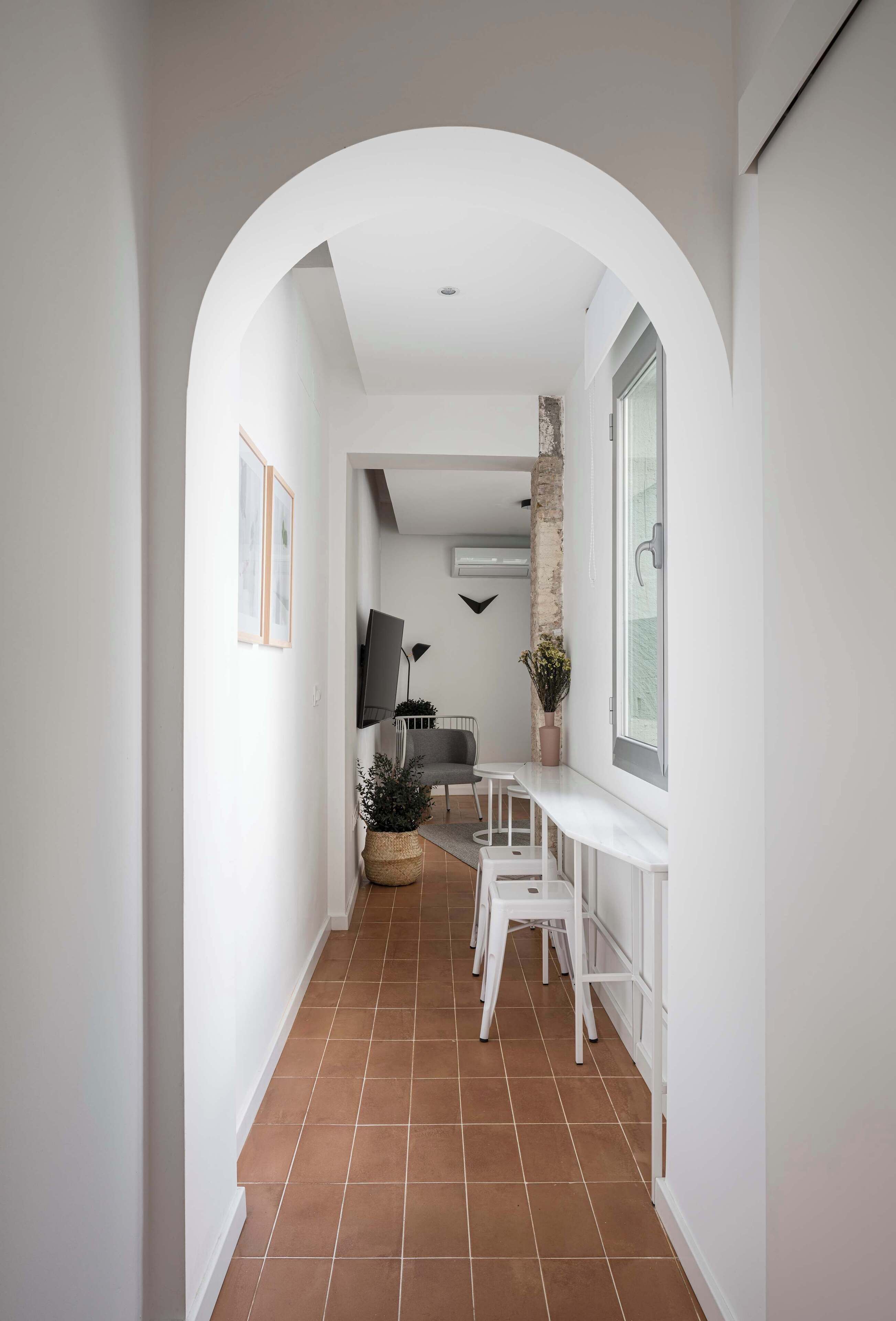 Un couloir étroit, surmonté d’une arche blanche, mène à un espace de vie. On y trouve des tableaux encadrés accrochés au mur, un téléviseur à écran plat et une plante en pot.