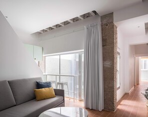 Ein modernes Wohnzimmer mit einem grauen Sofa, einem gelben Kissen, einem Glastisch und einem großen Fenster mit Vorhang.