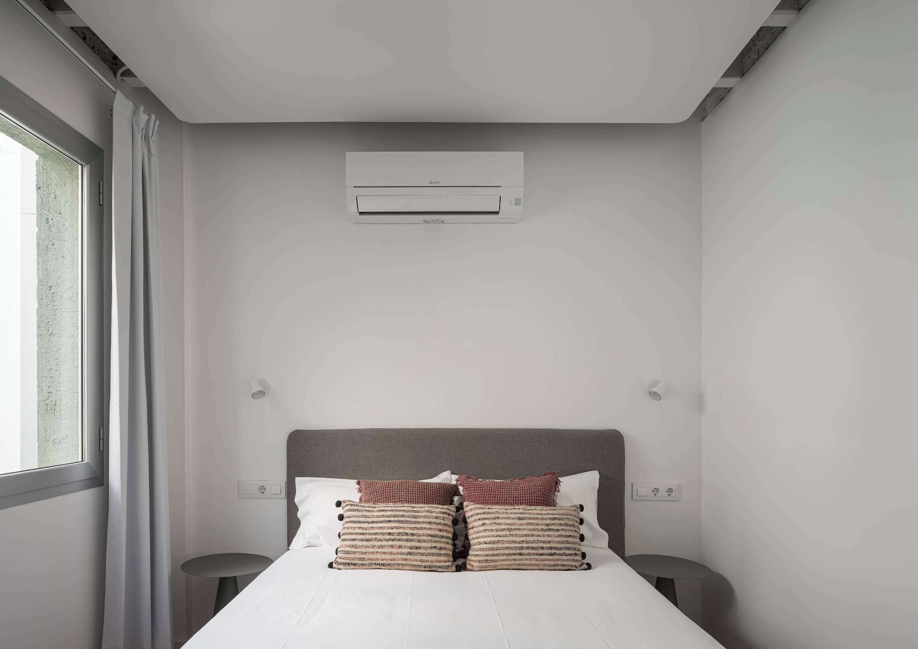 Quarto de hotel moderno com uma cama, duas mesas de cabeceira, uma janela com cortinas e um aparelho de ar condicionado na parede.
