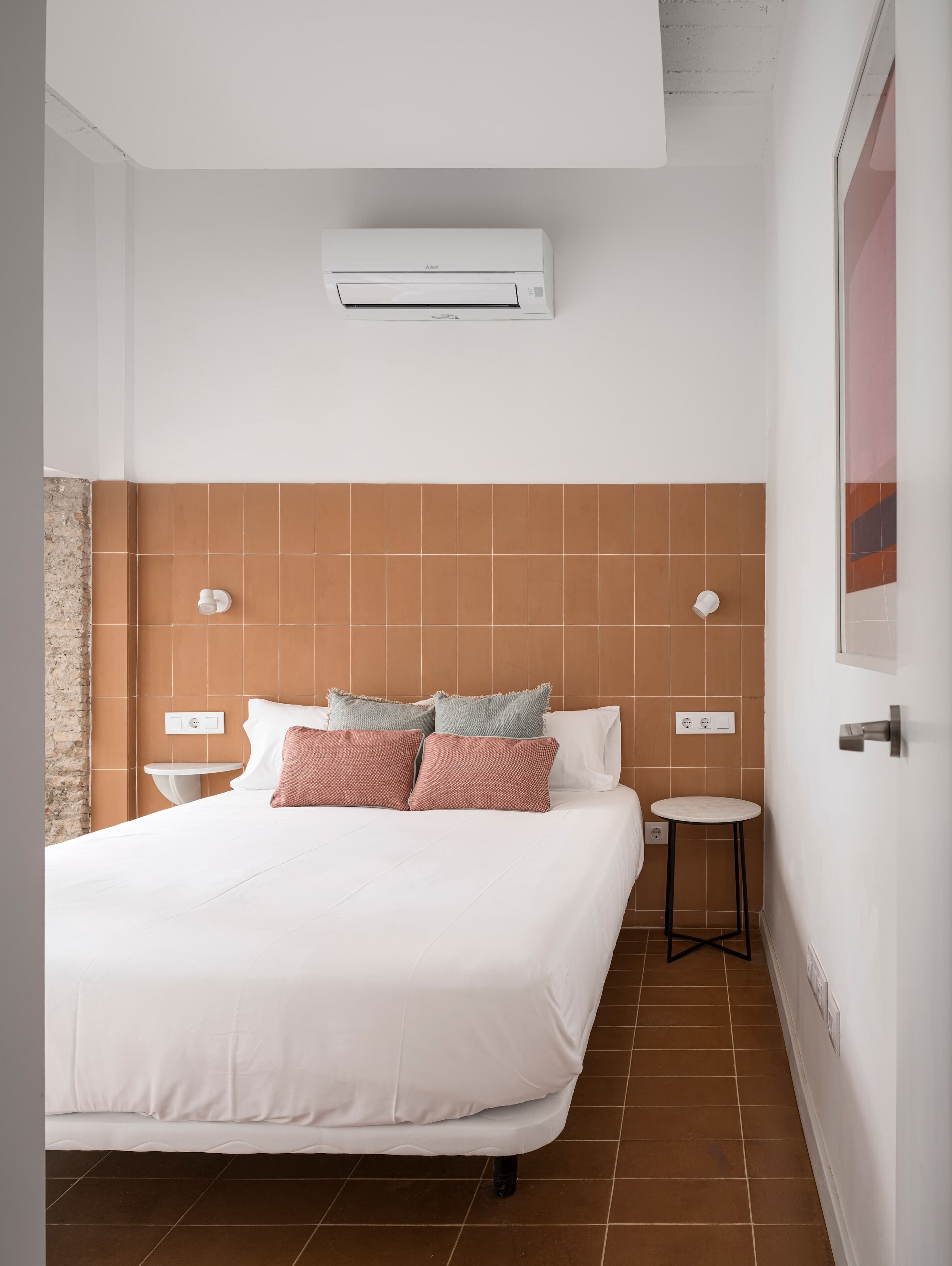 A hotel room with a large bed, a small bedside table, a wall-mounted air conditioner, and a tiled wall.