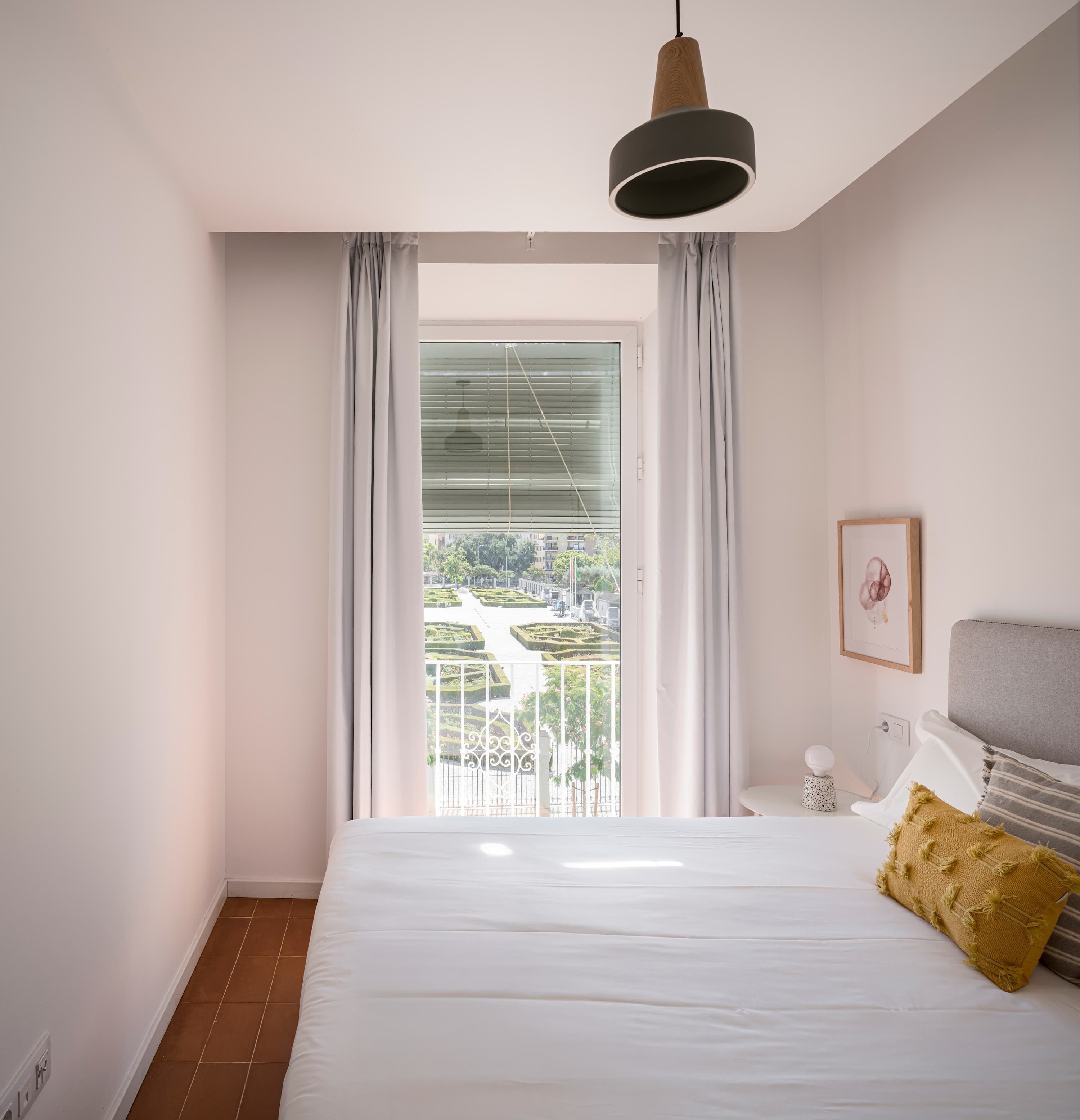Une chambre avec un grand lit, une table de chevet, un luminaire suspendu, une fenêtre avec des rideaux et une vue sur un jardin et des bâtiments à l’extérieur.