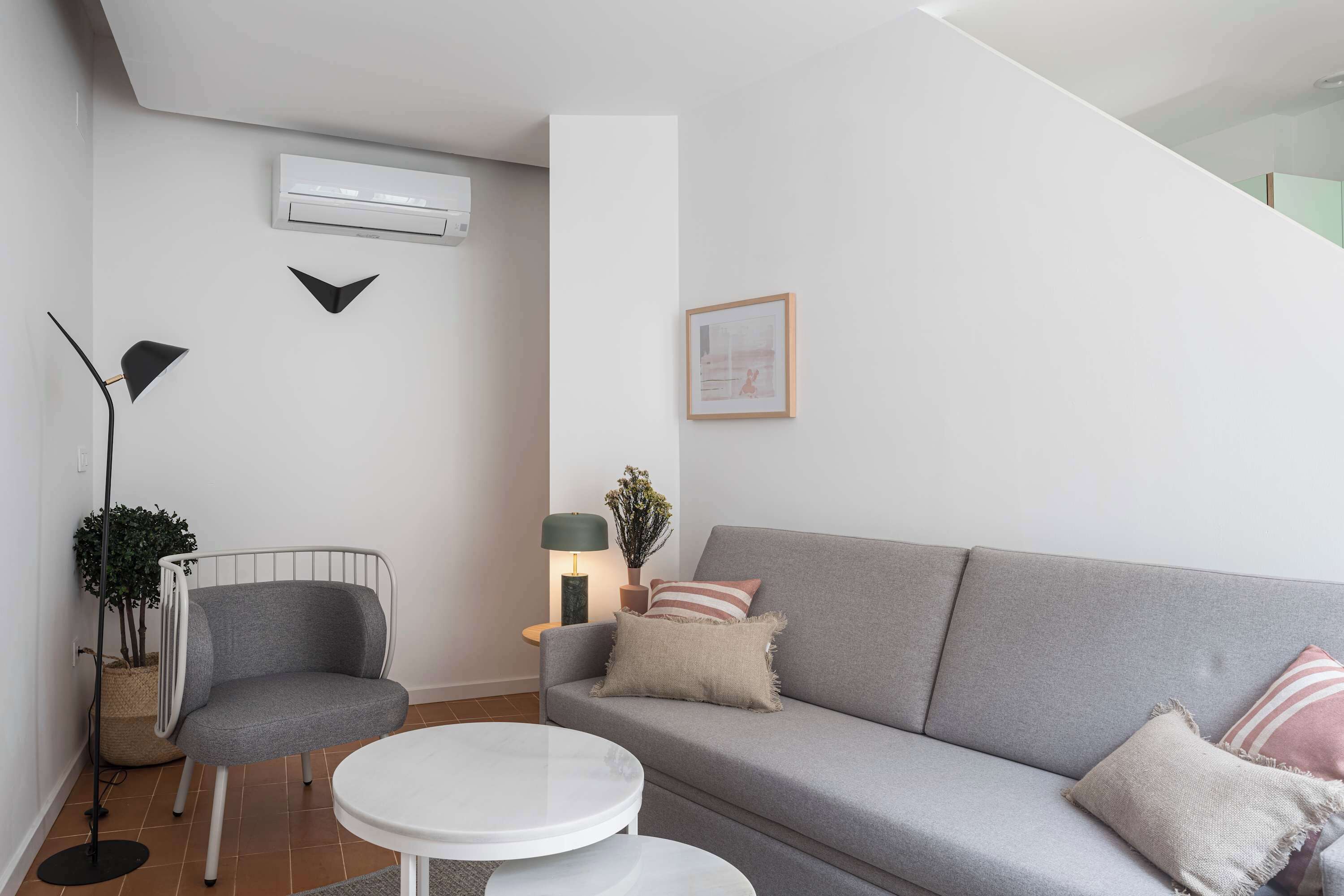 Ein modernes Wohnzimmer mit einem grauen Sofa, einem weißen runden Tisch, einer Stehlampe und einem kleinen Beistelltisch mit Lampe und Pflanze.