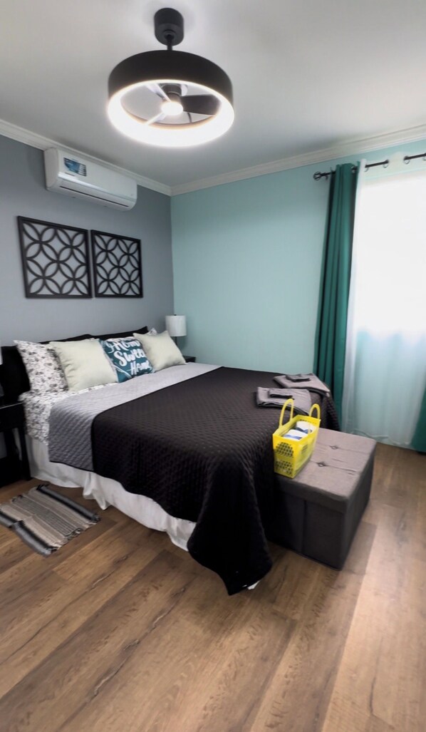A bedroom with a bed, bedside table, lamp, air conditioning unit, decorative wall panels, and a window with curtains.