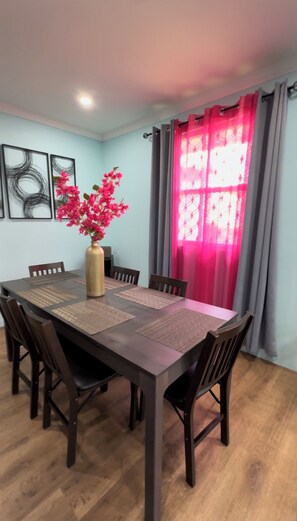 A dining area with a rectangular table, six chairs, a vase with pink flowers, and a window with pink curtains.