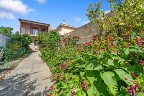 A pathway leading to a building with a balcony, surrounded by lush greenery and flowering plants.