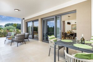 Un espace de restauration extérieur moderne, doté d’une terrasse fermée par des baies vitrées, de meubles en osier et d’une table dressée pour deux personnes, avec une bouteille de vin.