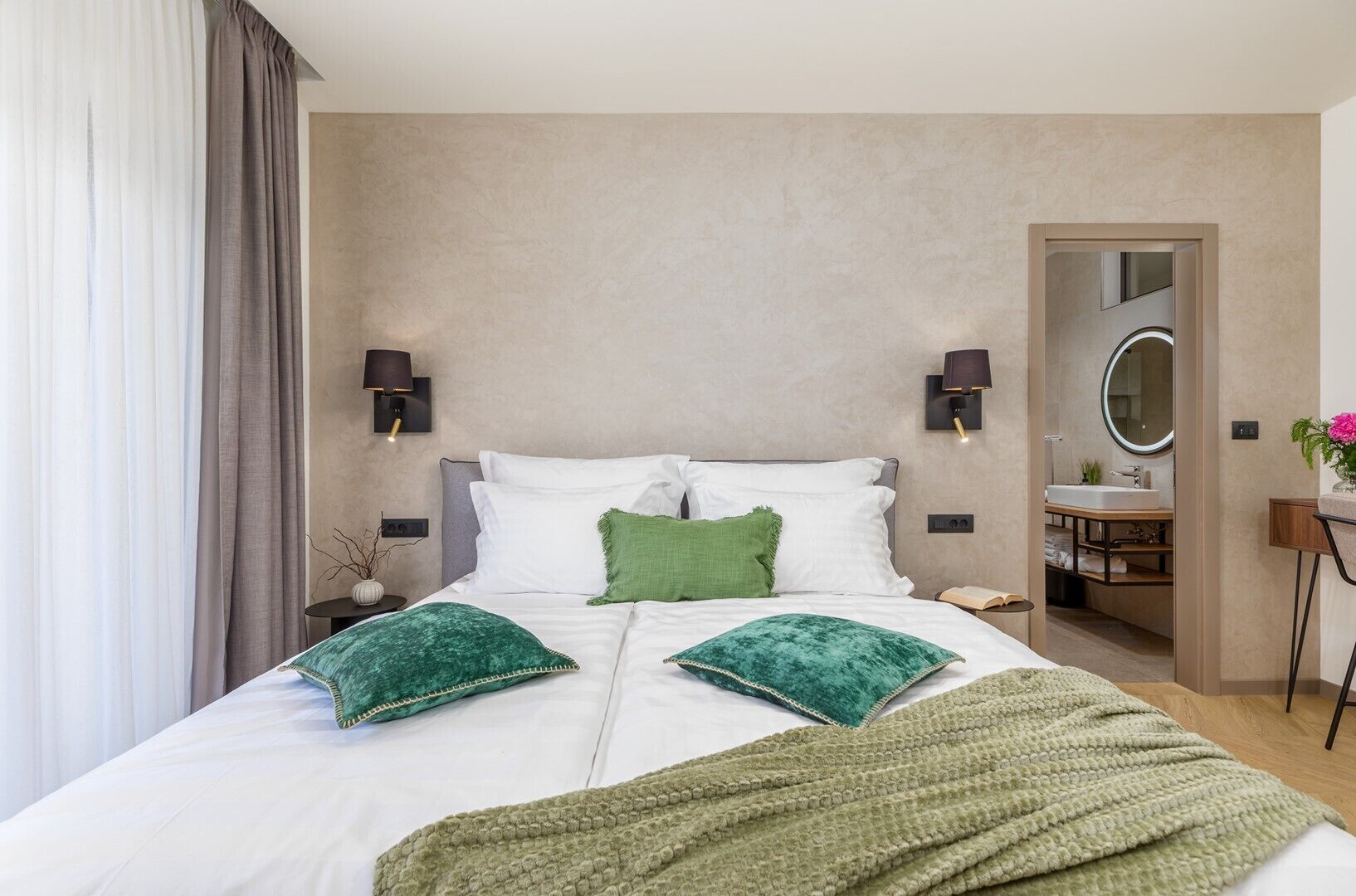 A hotel room with a bed, two wall-mounted lamps, a bedside table, a mirror, and a bathroom visible through an open door.