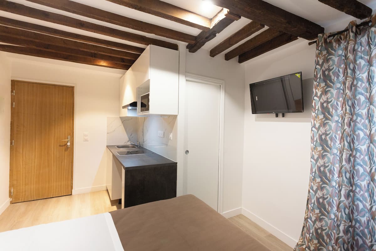 Une petite pièce comprenant un lit, un coin cuisine, une porte en bois et un téléviseur fixé au mur.