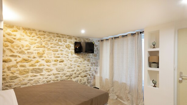 Une chambre avec un mur en pierre, un lit, un téléviseur fixé au mur et une porte surmontée d’une étagère.