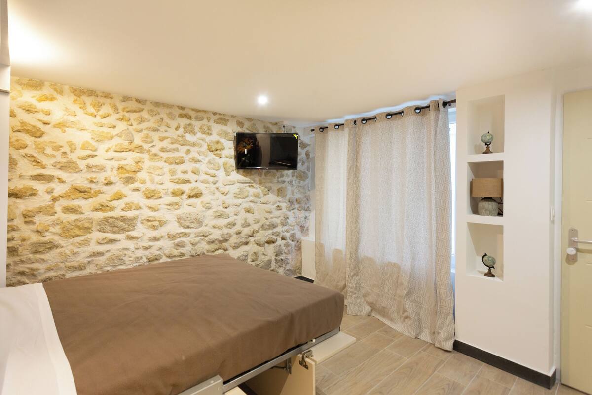 Une chambre avec un mur en pierre, un lit, un téléviseur fixé au mur et une porte surmontée d’une étagère.