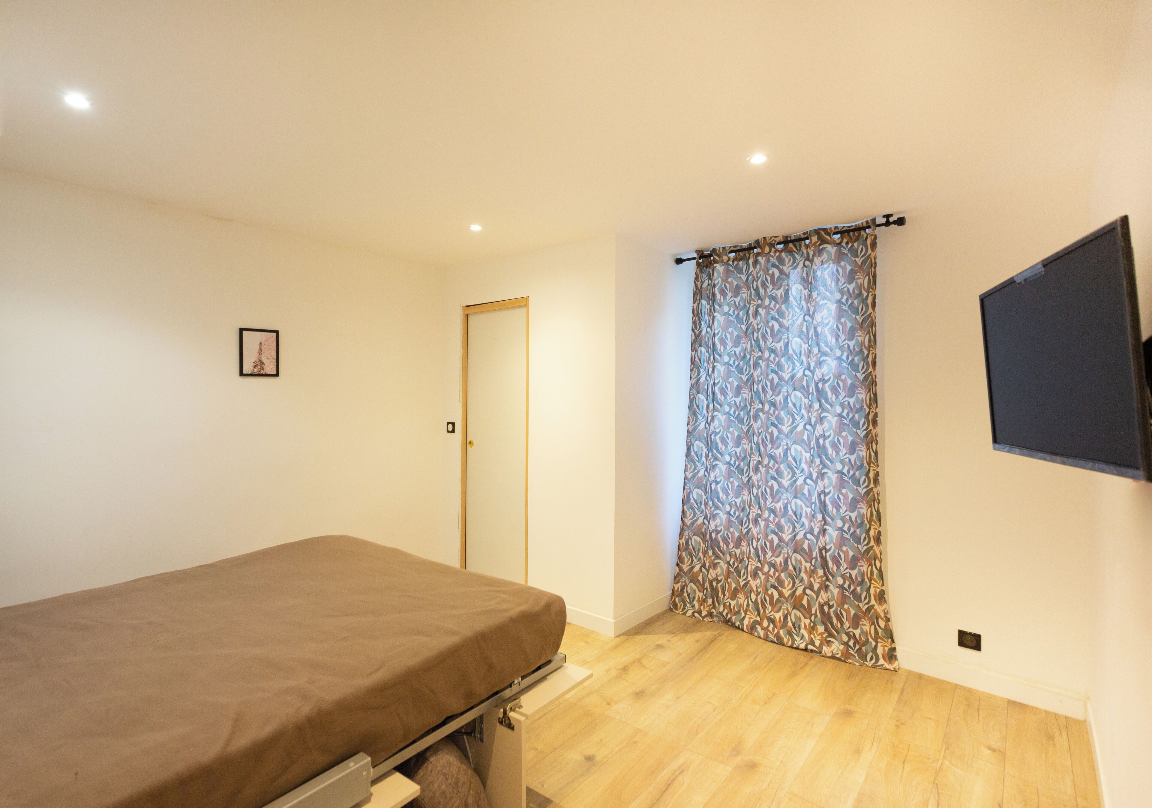 A bedroom with a bed, a door, a patterned curtain, and a mounted TV.