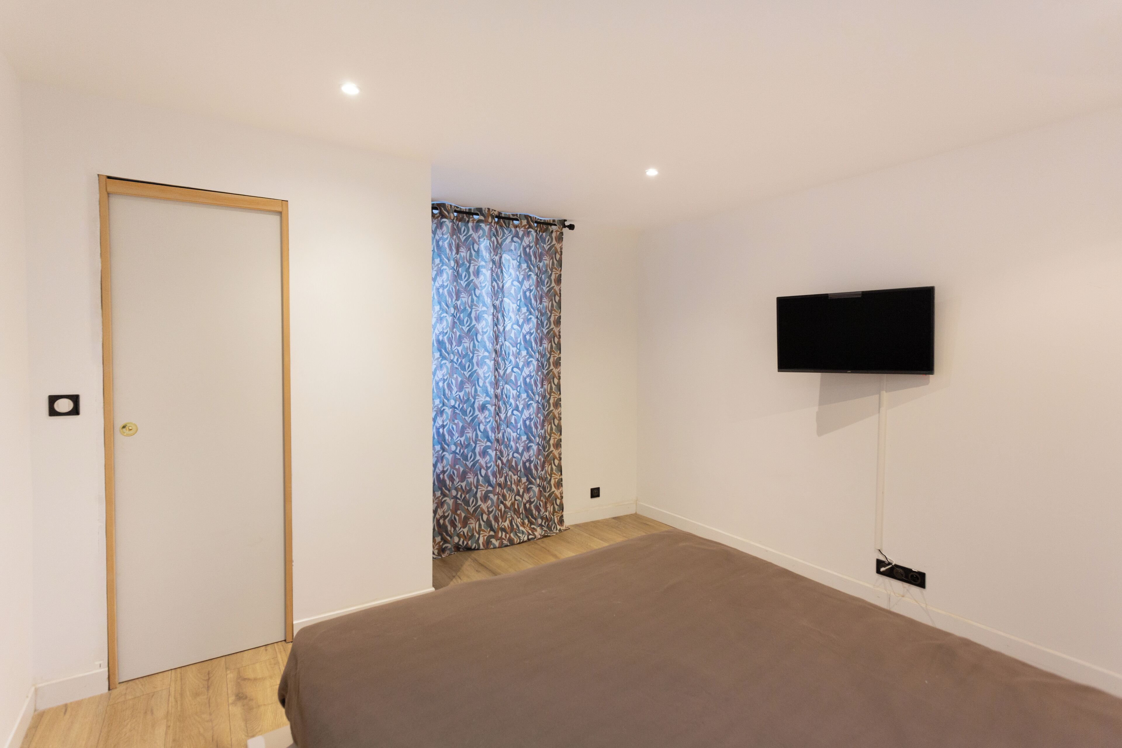 Un dormitorio con una cama, televisor montado en la pared, una puerta y cortina estampada.