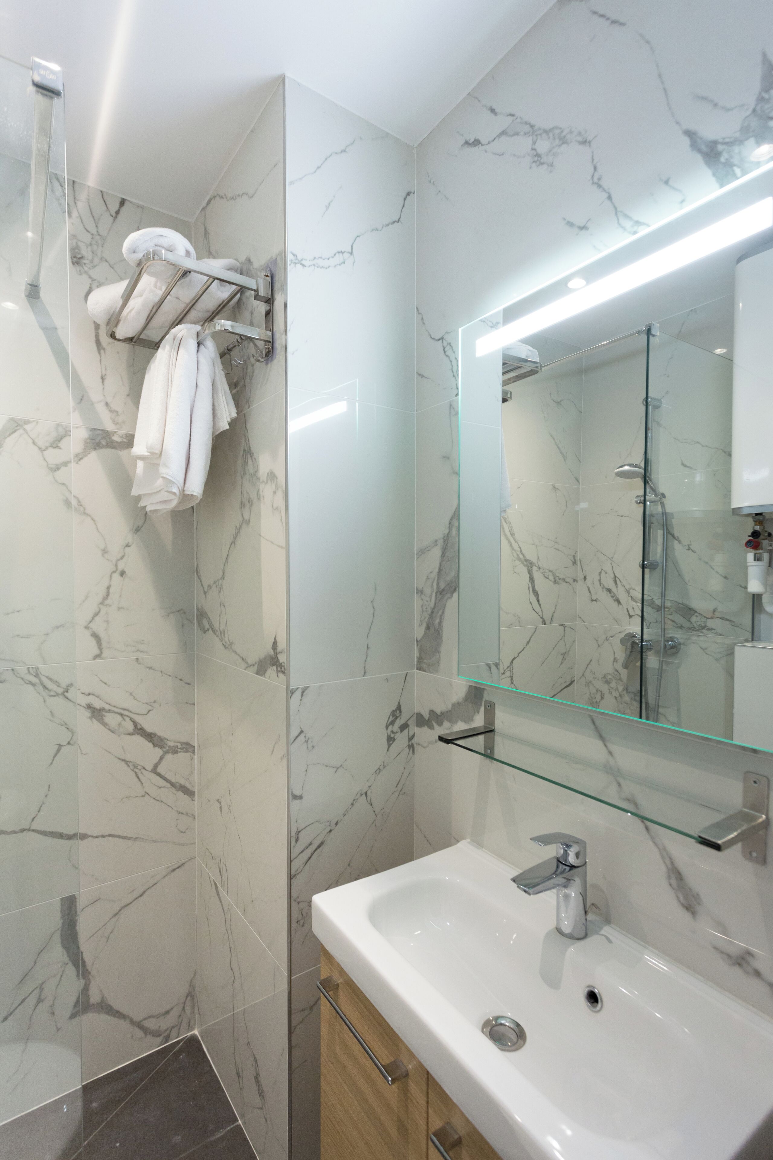 A modern bathroom with marble walls, a glass shower enclosure, a white sink, and a towel rack.