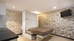 Une chambre à coucher de taille réduite, comprenant un lit, des rangements intégrés, un mur en pierre et un téléviseur fixé au mur.