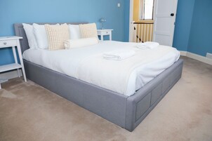 Un lit bien fait, avec du linge de lit blanc et une tête de lit grise, dans une pièce aux murs bleus et au tapis beige.