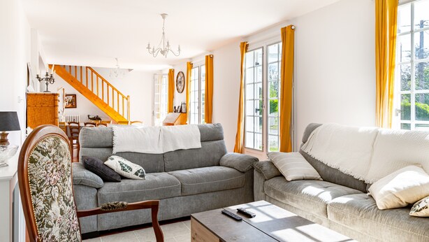 A living room with a grey sofa, a wooden chair, a coffee table, and a staircase with yellow curtains.
