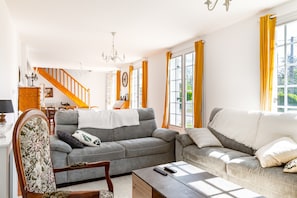 Uma sala de estar com um sofá cinza, uma cadeira de madeira, uma mesa de centro e uma escada com cortinas amarelas.