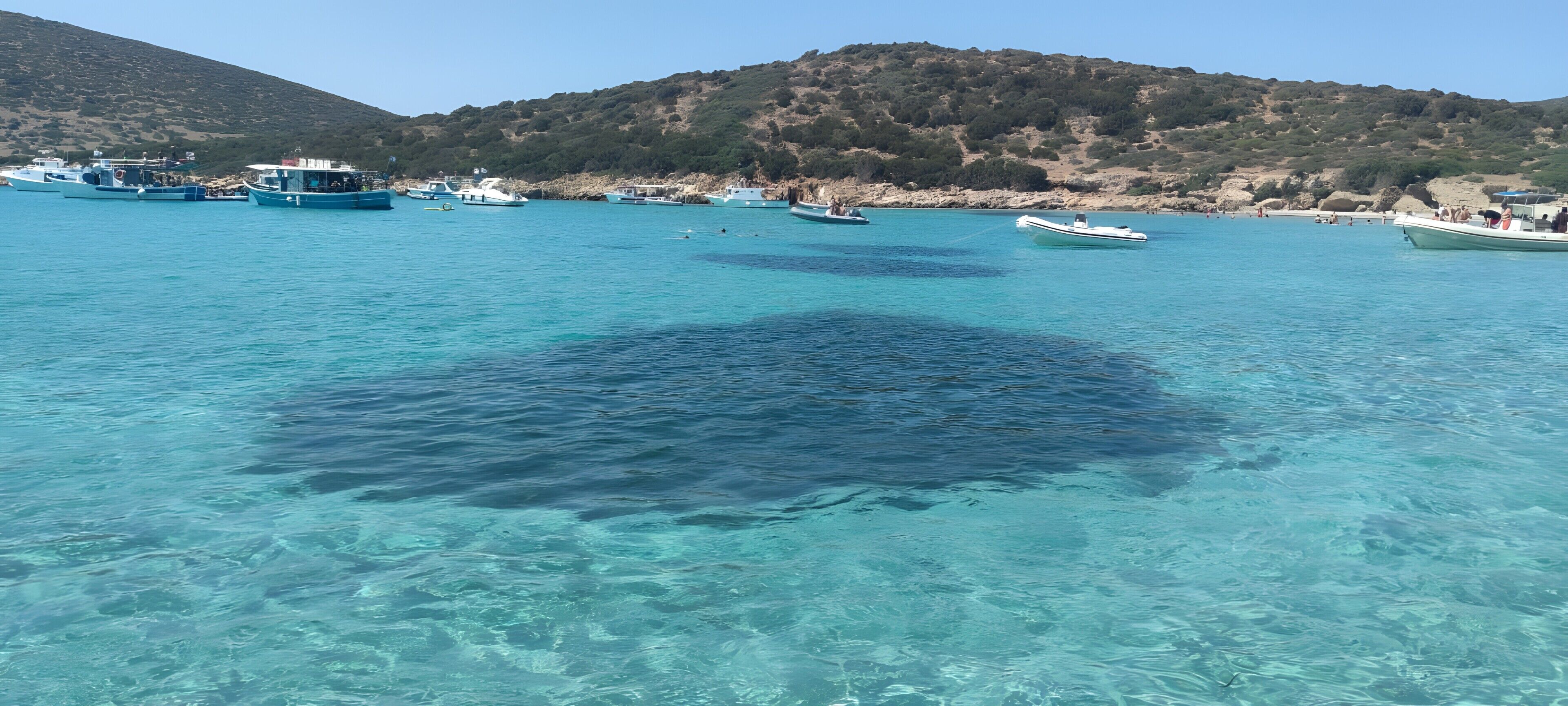 Zona costera con barcos, agua cristalina y un paisaje montañoso al fondo.