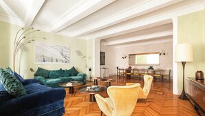 Ein geräumiges Wohnzimmer mit einem blauen Sofa, einem gelben Sessel und einem Fischgrätparkett. Im Hintergrund ist ein Essbereich mit Tisch und Stühlen zu sehen.