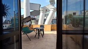A balcony with a green hammock, a small table, and a chair. There are large white columns and a building with a modern design in the background. - 3-bedroom apartment in Batumi (Batumi)