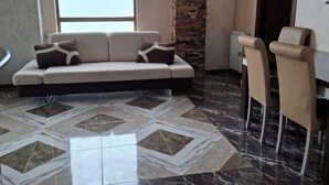 A modern living room with a sectional sofa, a dining area with two chairs, a large window, and a stone accent wall. - 3-bedroom apartment in Batumi (Batumi)
