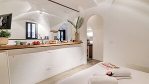 A modern hotel room with a kitchen area, a dining table, and a bed with towels.