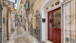 A narrow cobblestone alley with a wooden door and metal gate, flanked by buildings with balconies and shutters.