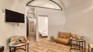 A modern living room with a herringbone floor, a flat-screen TV mounted on the wall, a brown sofa, two round tables with breakfast items, and a door leading to a bedroom. - Boutique Apartment Casa Del Barone (Ventimiglia)